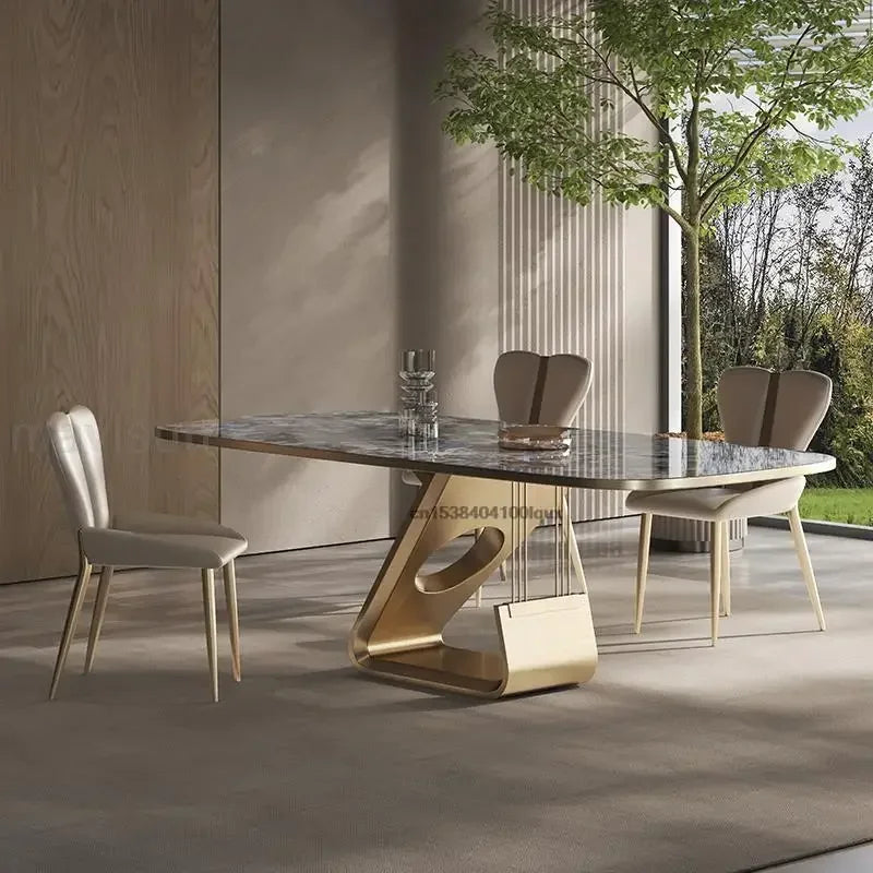 Modern dining table with marble top and gold base in a room with large windows.