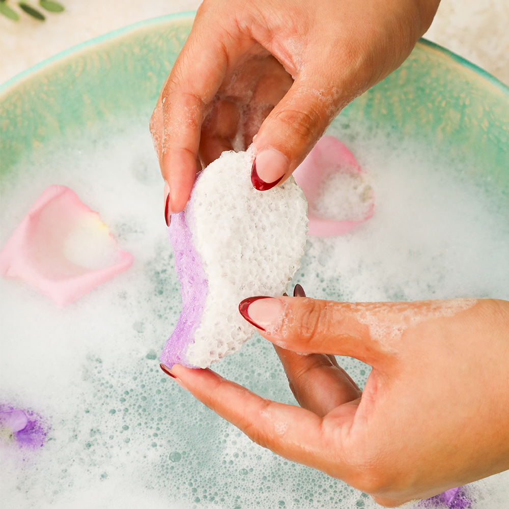Hand holding a white and purple bath bomb with a soapy water background