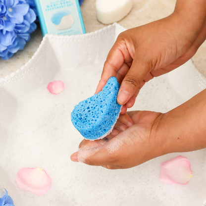 Hand holding a blue sponge with soap bubbles, surrounded by flowers and bath products.