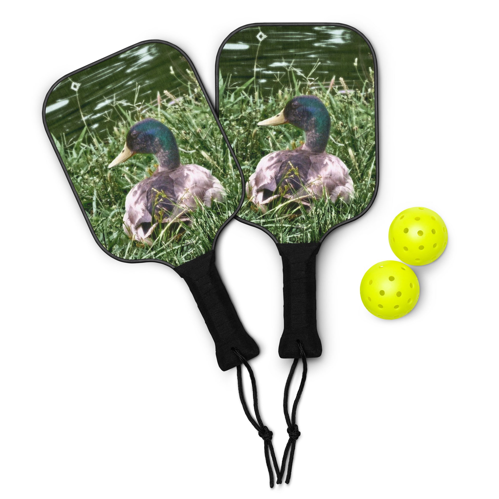 Two pickleball paddles with duck design on grass and two yellow pickleballs.